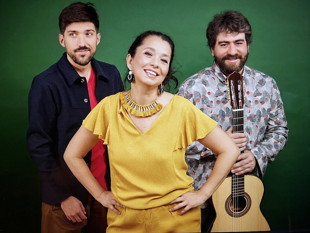 Céline Rudolph steht lächelnd vor Henrique Gomide und João Luís Nogueira mit Akustikgitarre.